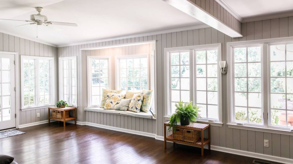 Bright sunroom with large windows, a cozy window seat, dark hardwood floors, gray walls, a ceiling fan, and minimalistic decor featuring plants.